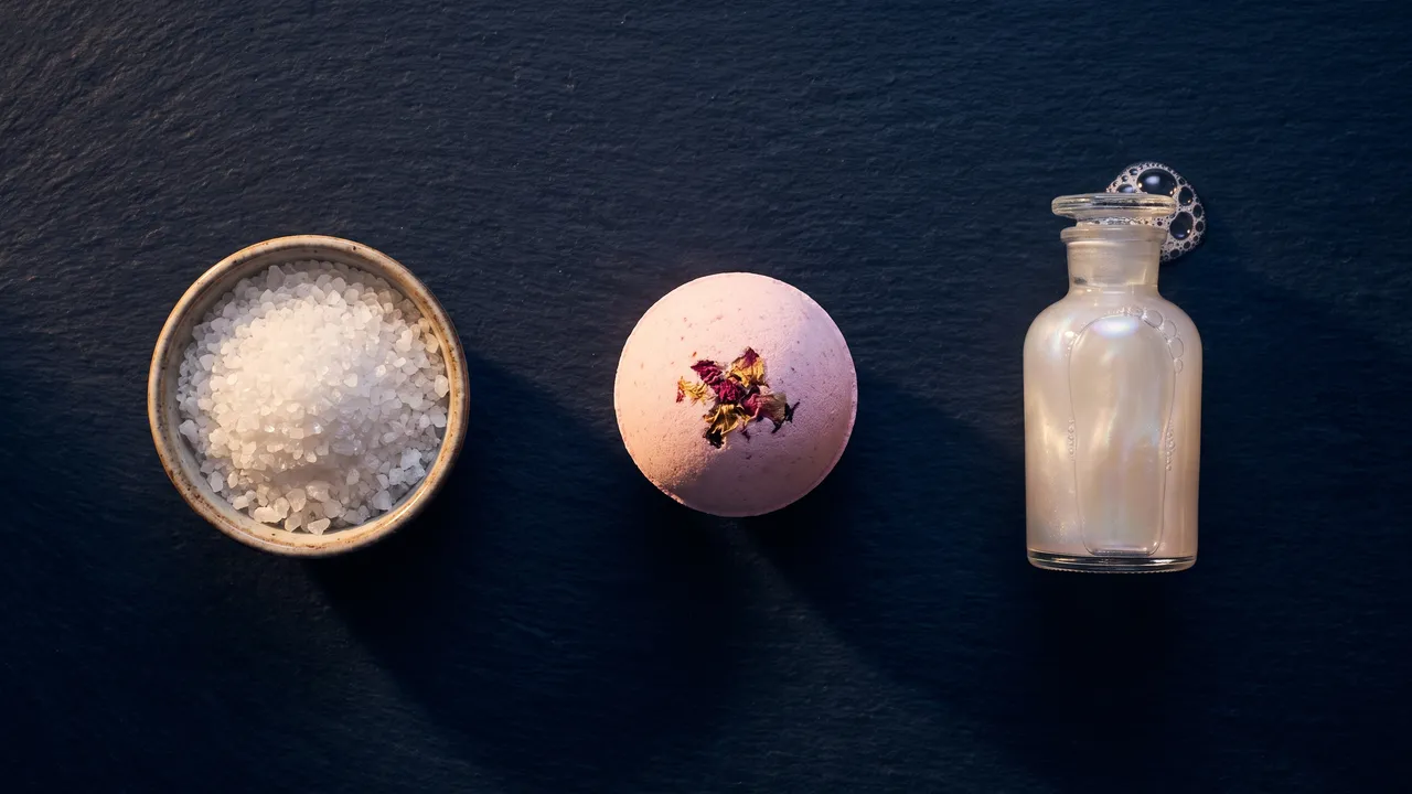 A dish of bath salts, a pink bath bomb and a bottle of bubble bath side by side on dark stone